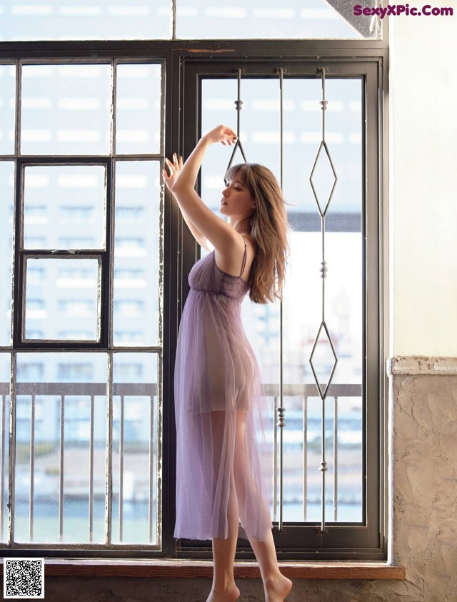 A woman in a purple dress standing by a window.